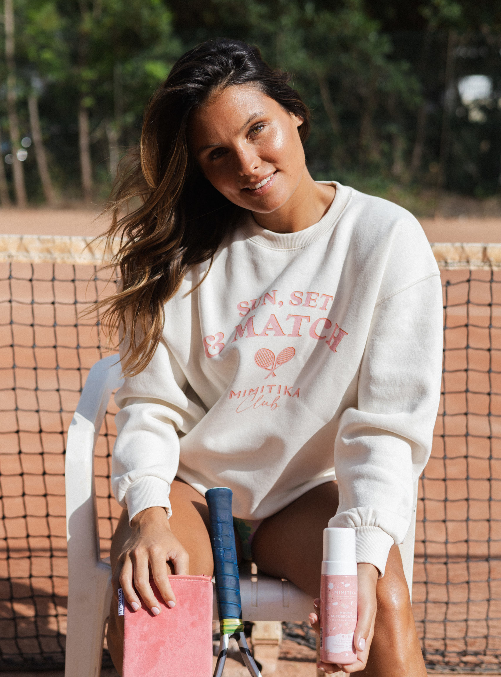 Une jeune femme sur un cours de tennis présente un produit de cosmétique. Elle est chatain et la peau mat. Elle a un maquillage très solaire et naturel.