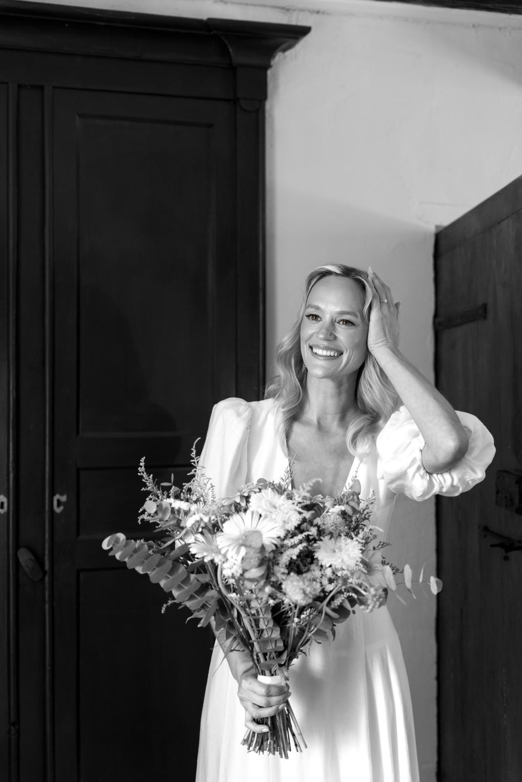 Une photo en noir et blanc d'une mariée blonde avec qui tient un très joli bouquet de fleurs. Elle porte une robe blanche en lin et elle un maquillage très naturel.